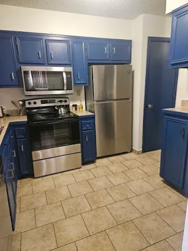 Blue cabinets, stainless steel appliances in a kitchen with beige tiled floor.