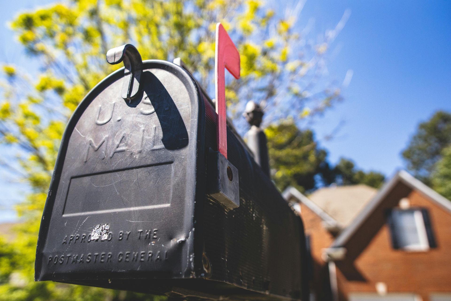 Black U.S. Mail mailbox with red flag up, set against a blurred house and blue sky.