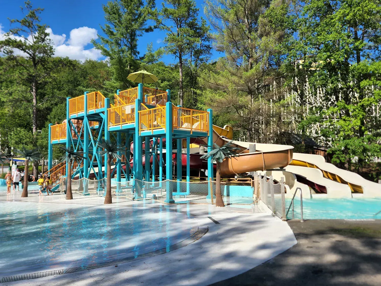Water park with teal structures, slides, and sprinklers, surrounded by trees.