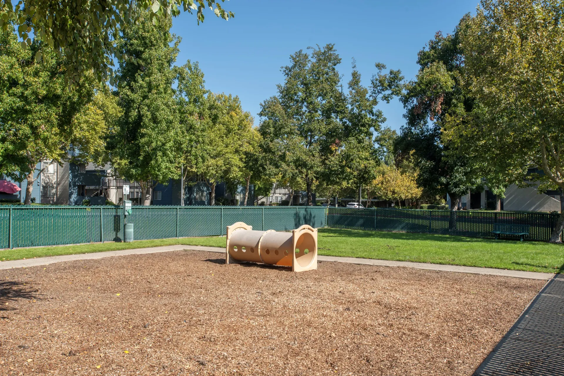 Playground with a tunnel-climber structure, wood-chip ground, and surrounding trees.