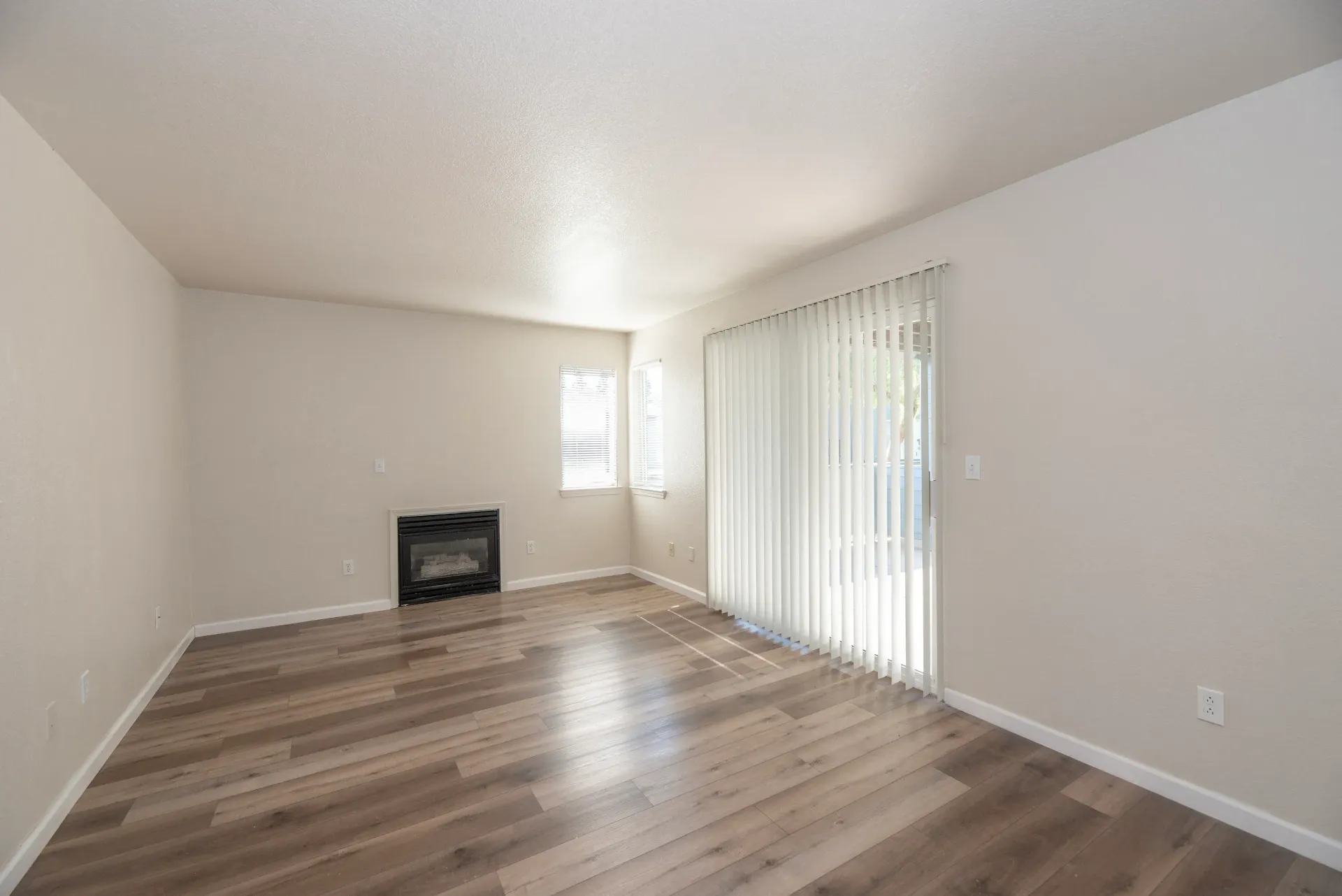 Living room with light walls, wood-look flooring, a fireplace on the far wall, and a sliding glass door with vertical blinds.