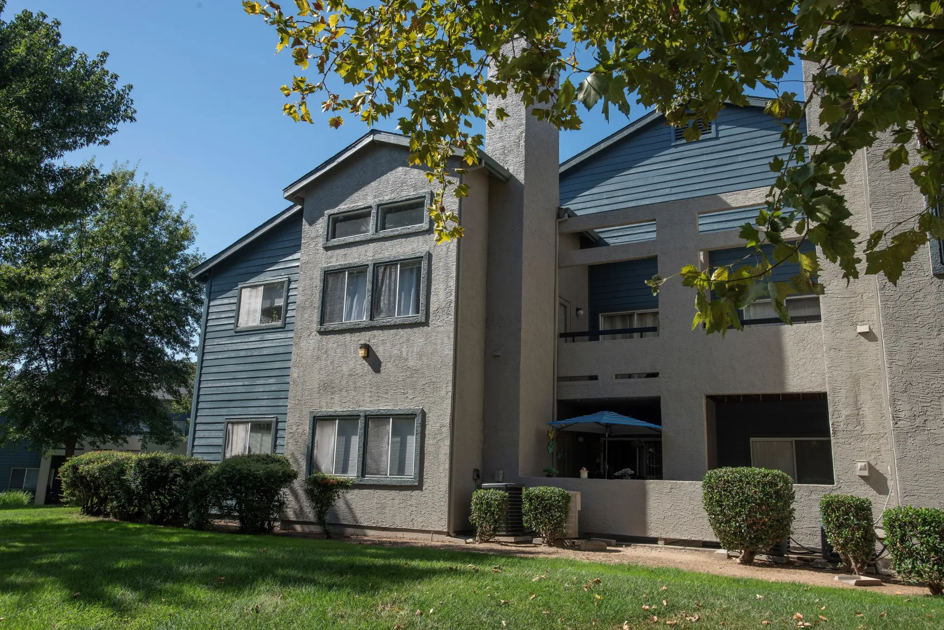 Exterior view of a two-story apartment building with beige stucco and blue siding, surrounded by green lawn and shrubs.