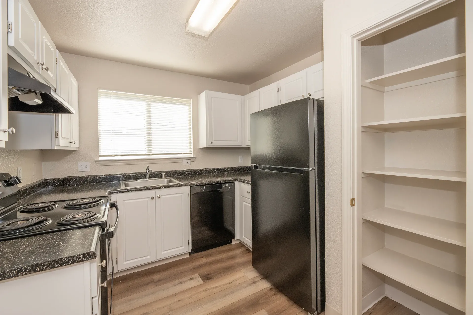 Kitchen in an apartment with white cabinets, black appliances, a window, and a pantry.