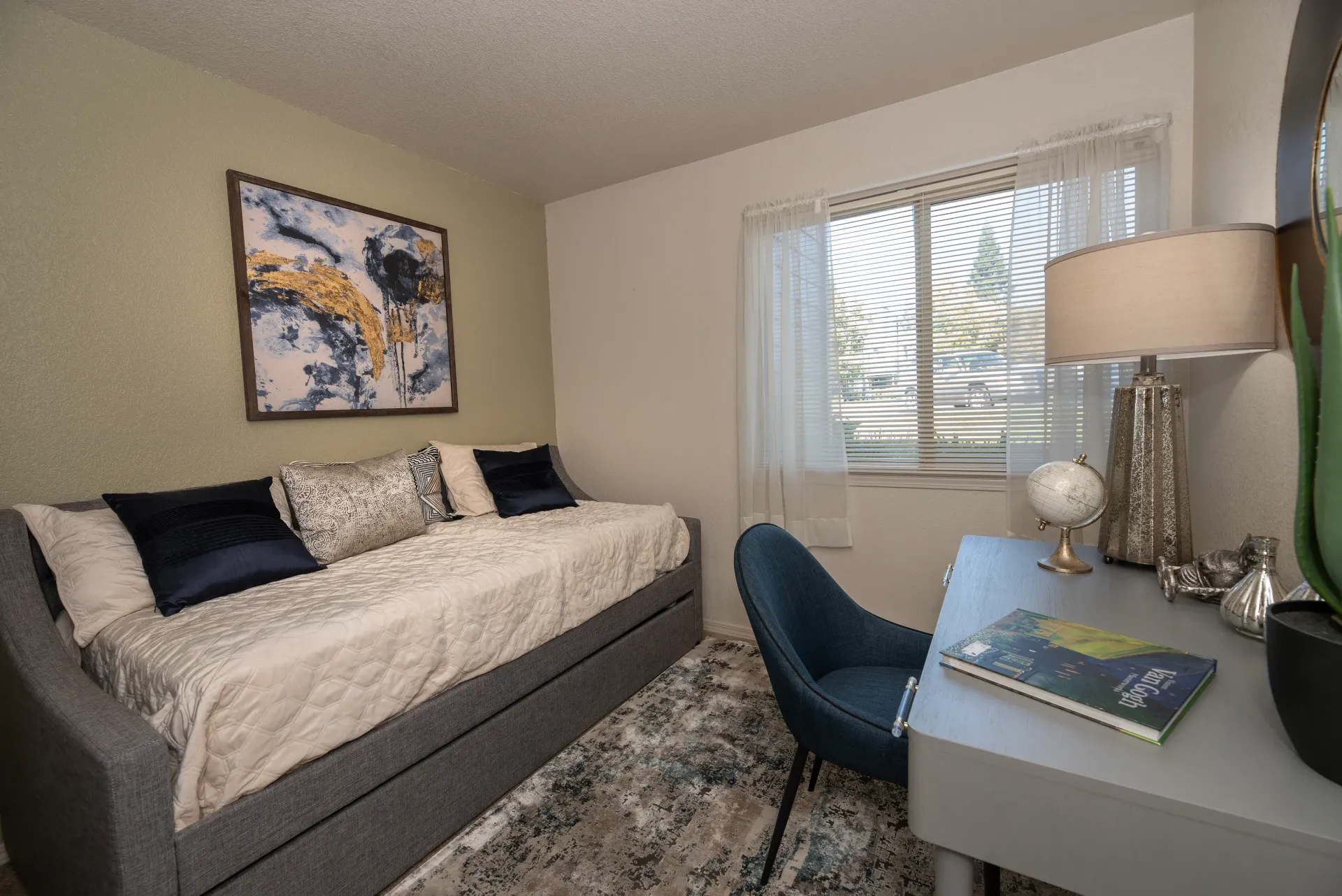 Bedroom area featuring a gray sofa bed, desk, chair, window with blinds, and wall art.