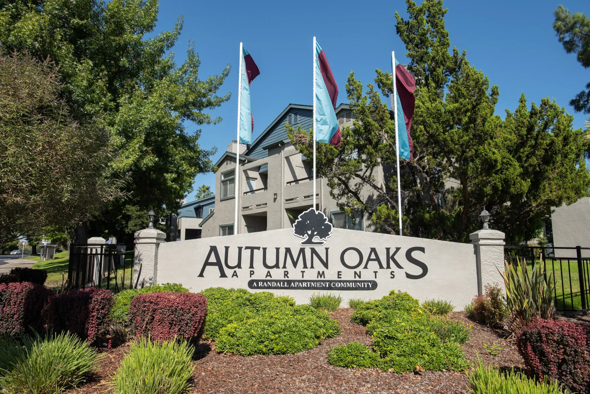 Exterior view of Autumn Oaks Apartments entrance with a large sign, flagpoles, and surrounding landscaping.