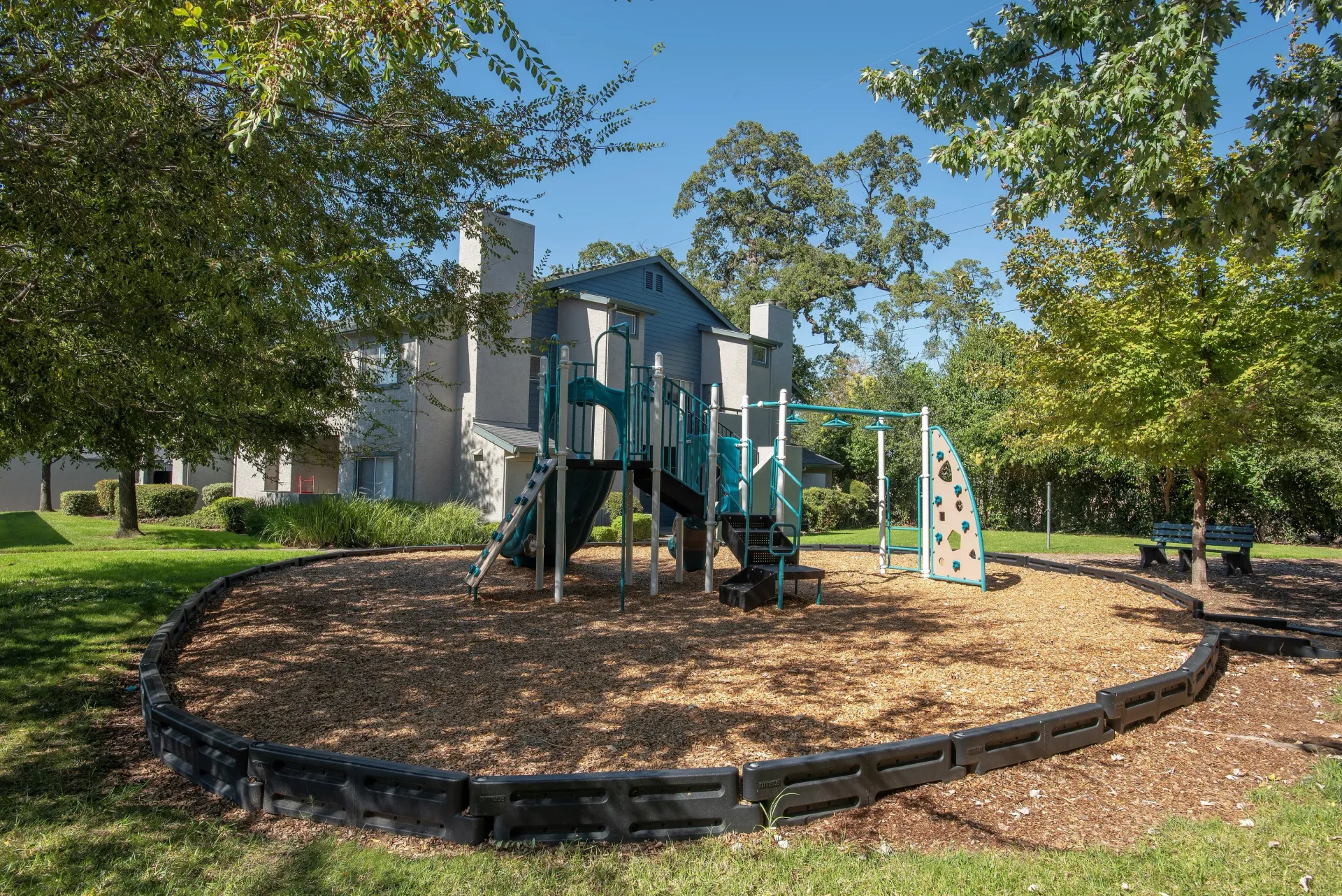 Playground with teal and white climbing structure, surrounded by mulch, in a courtyard beside an apartment building.