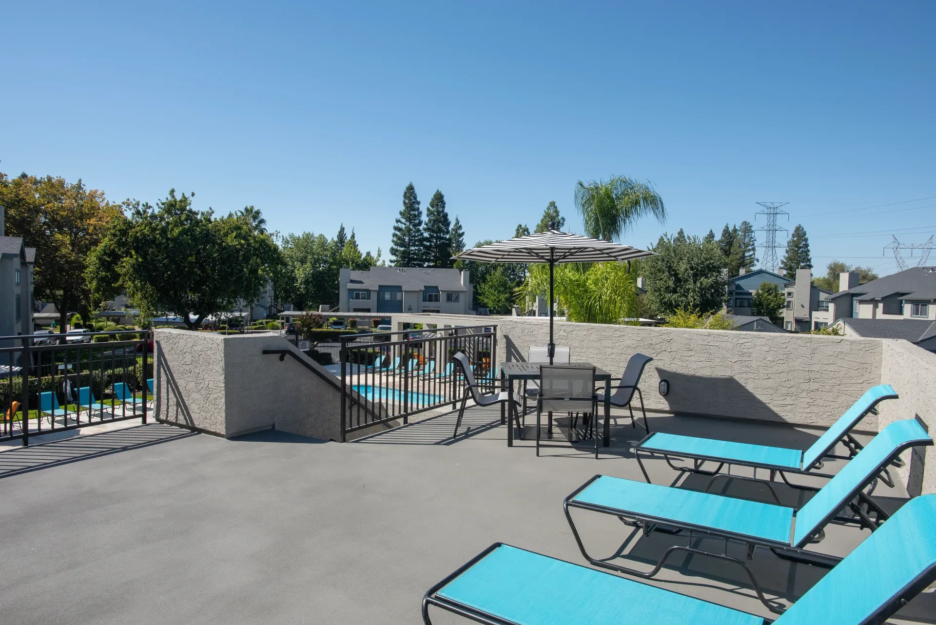 Rooftop communal patio with blue lounge chairs, a table, and umbrella overlooking a pool.