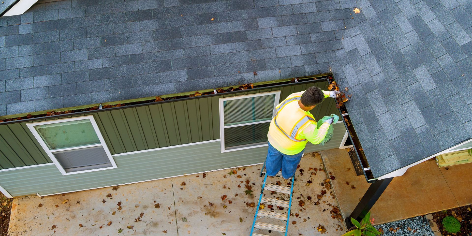 A person in a safety vest cleans leaves from a gutter on a house roof with a ladder.