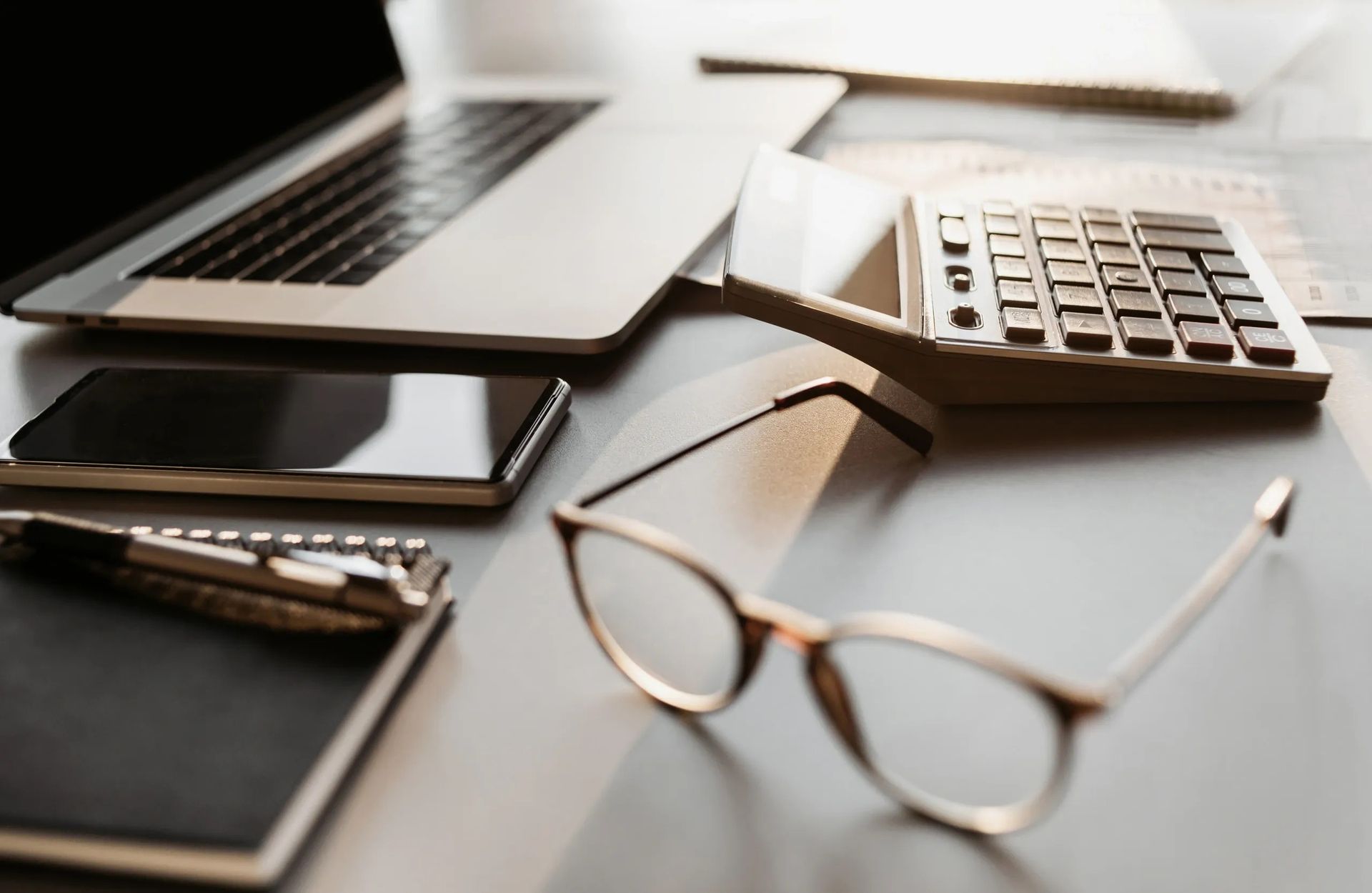 A pair of glasses is sitting on a desk next to a laptop and calculator.