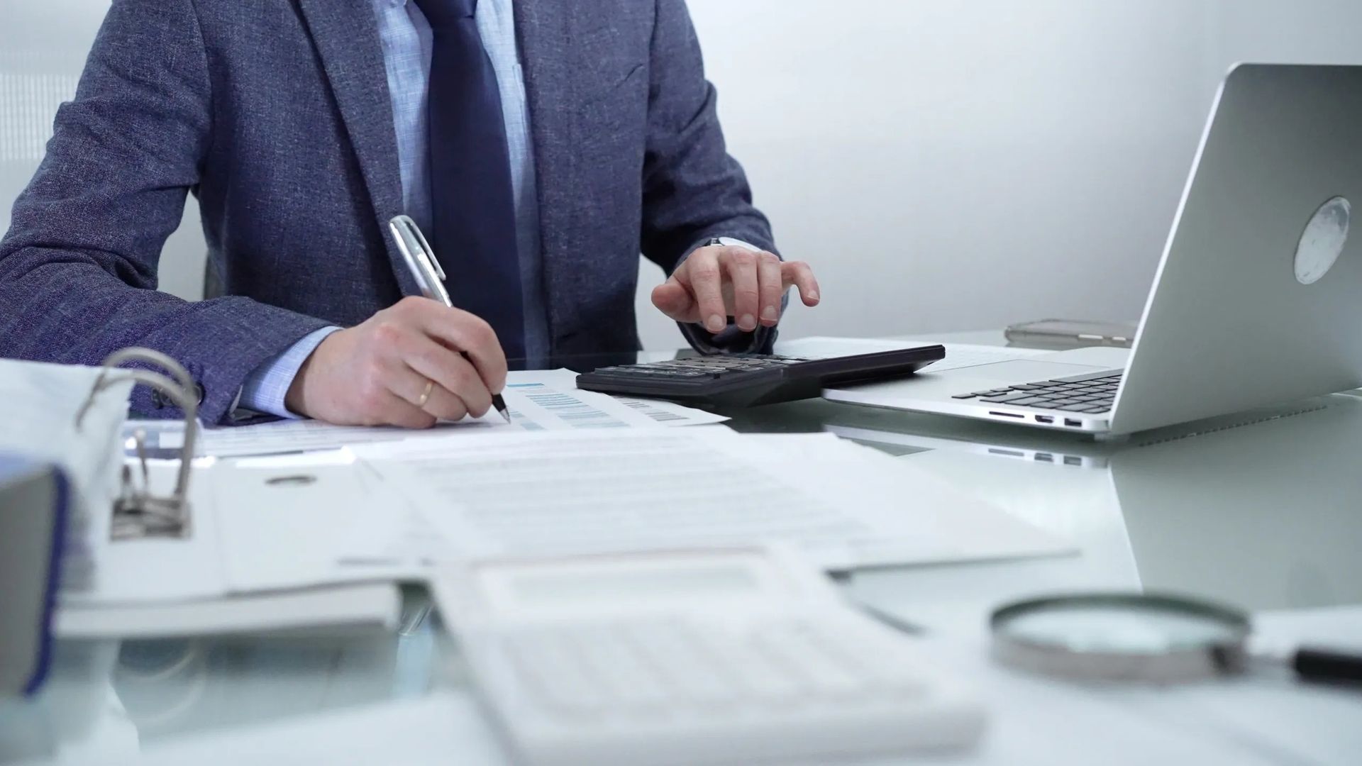 A man in a suit and tie is sitting at a desk using a laptop and a calculator.