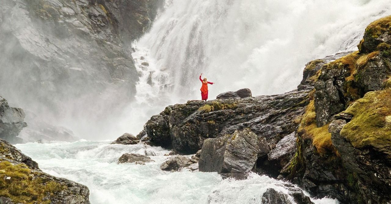 Een persoon in een rode jurk staat op een rots bij een waterval.