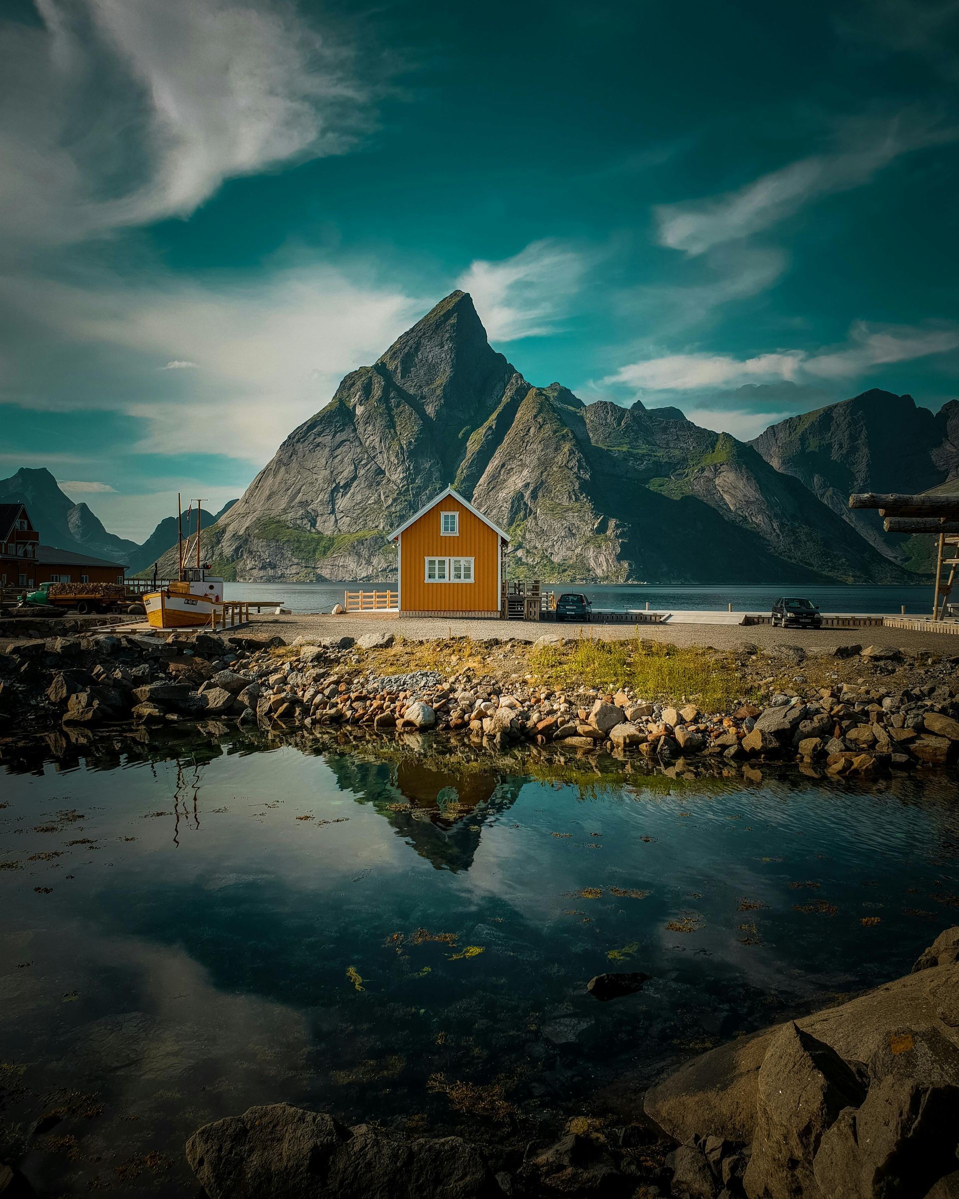 Een klein huisje staat aan de oever van een fjord tegenover een berg.