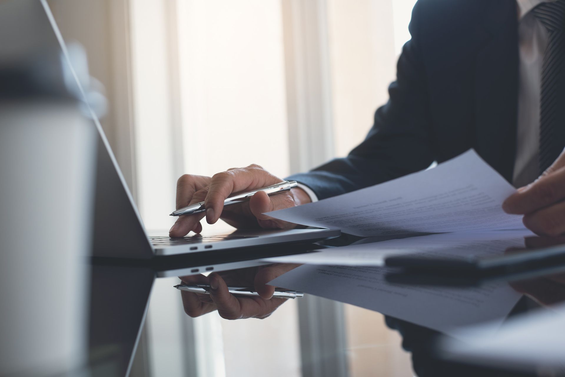 A man in a suit is using a laptop computer while holding a pen and a piece of paper.
