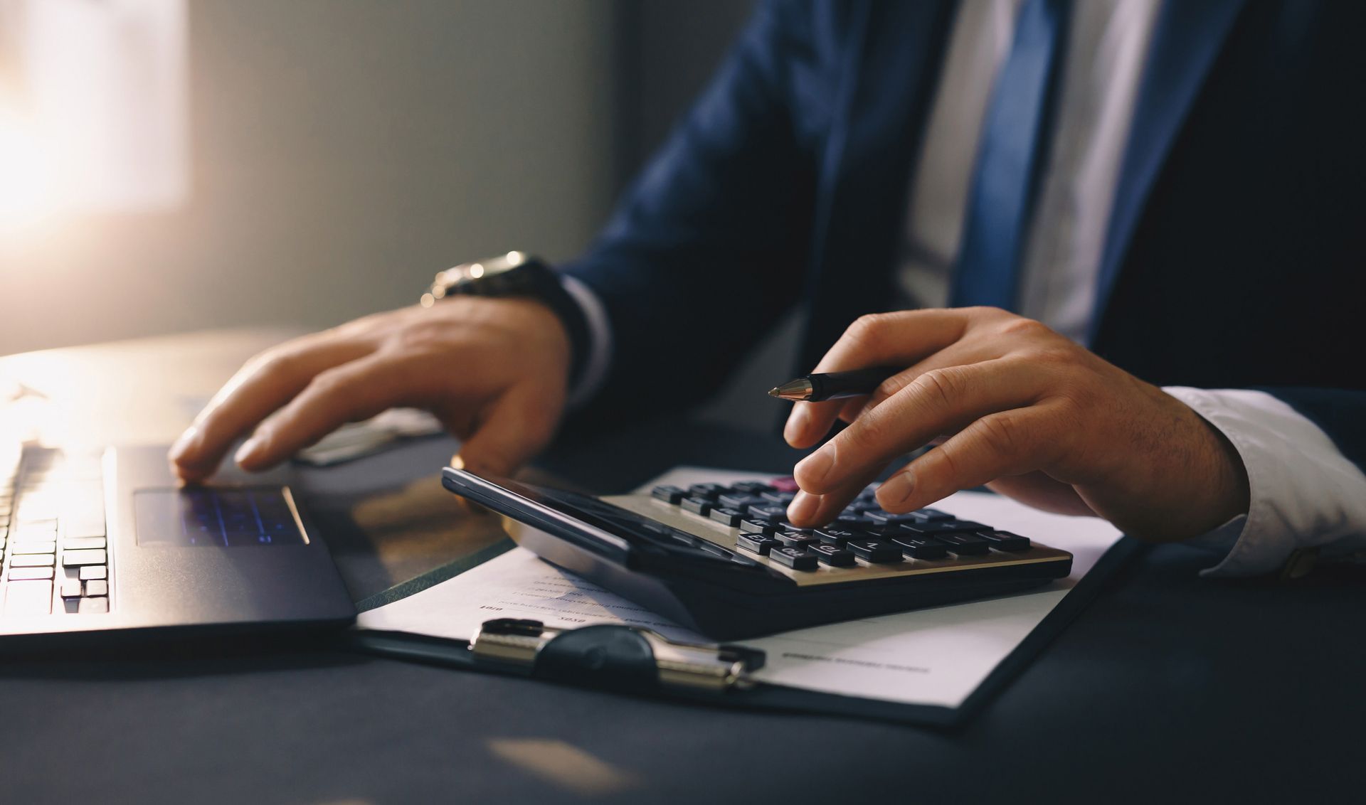 A man in a suit and tie is using a calculator and a laptop.