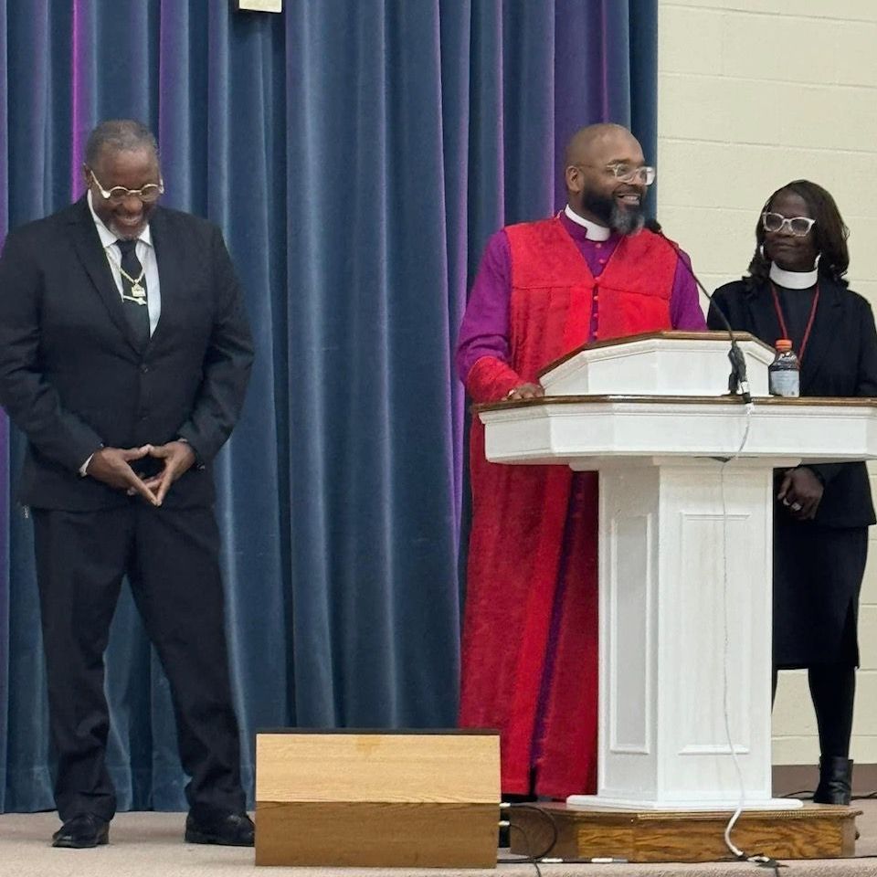 Three people at a podium. Man in suit smiles. Man in robe speaks. Woman in black stands by.