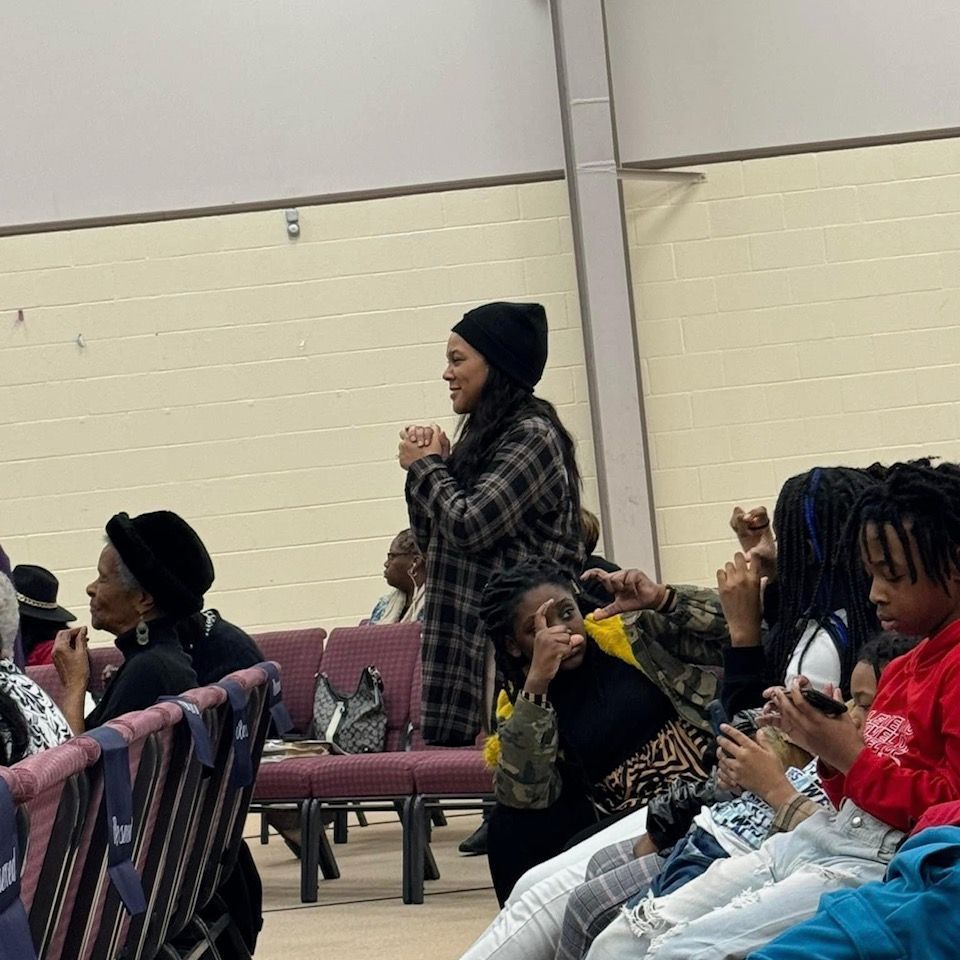Woman in plaid coat speaks at an event, people in the audience listen and react.