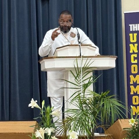 Man in white suit speaking at podium, blue backdrop, greenery, 