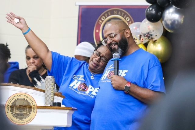 Two people in blue shirts embracing at a HOPE event. One person holding a microphone.