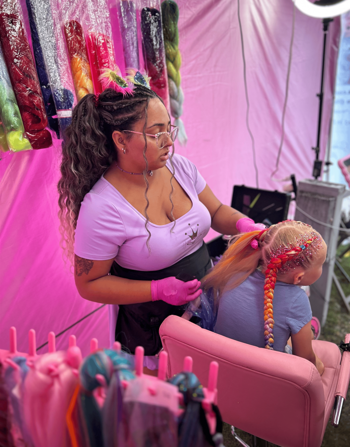 Hair stylist braiding a child’s hair in a pink salon booth, with colorful wigs hanging behind them.