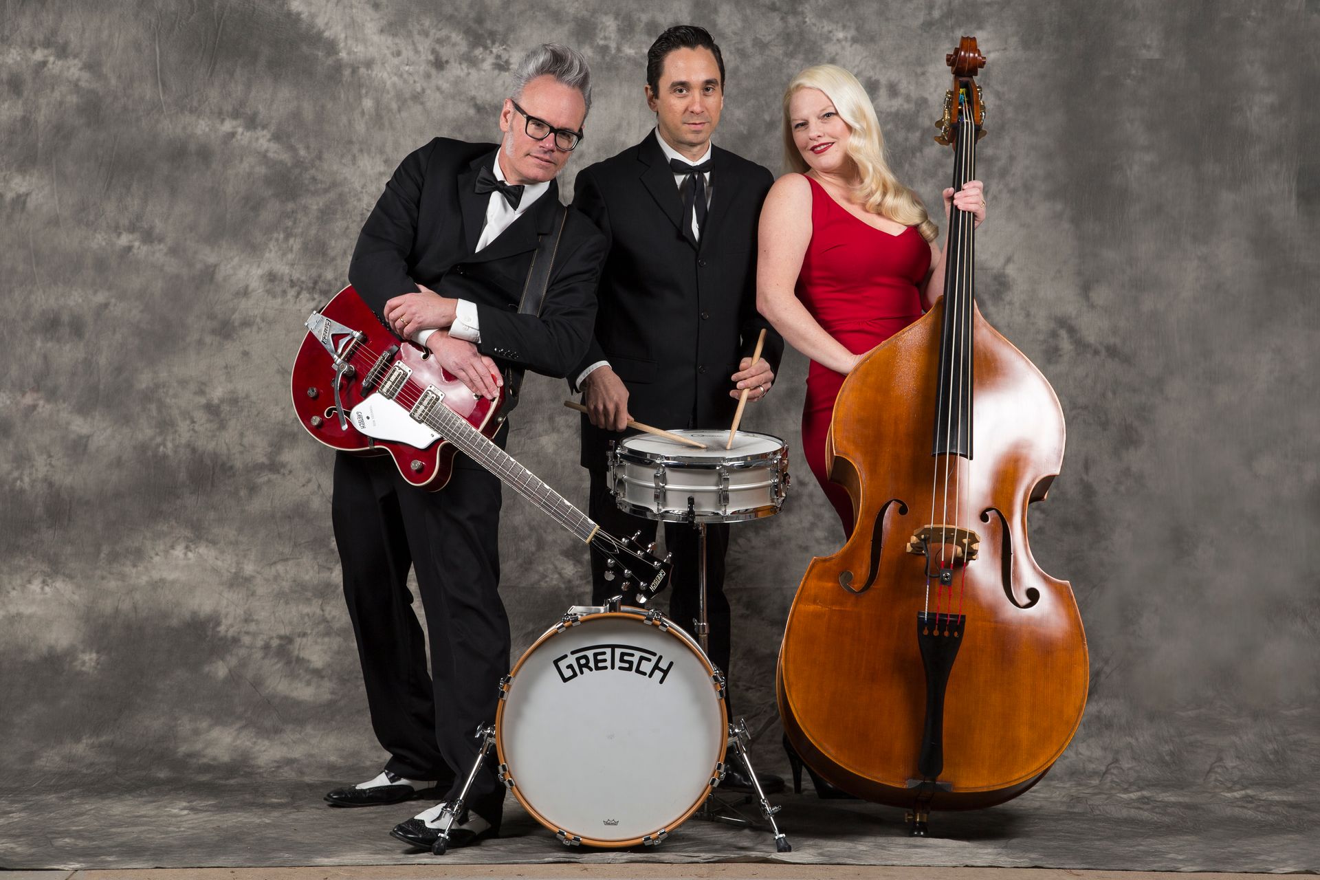 Three musicians pose in a studio with a red guitar, drum kit, and upright bass.