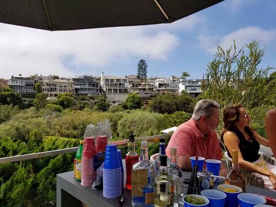 Outdoor patio with drinks on a table, overlooking greenery and white houses under a bright sky