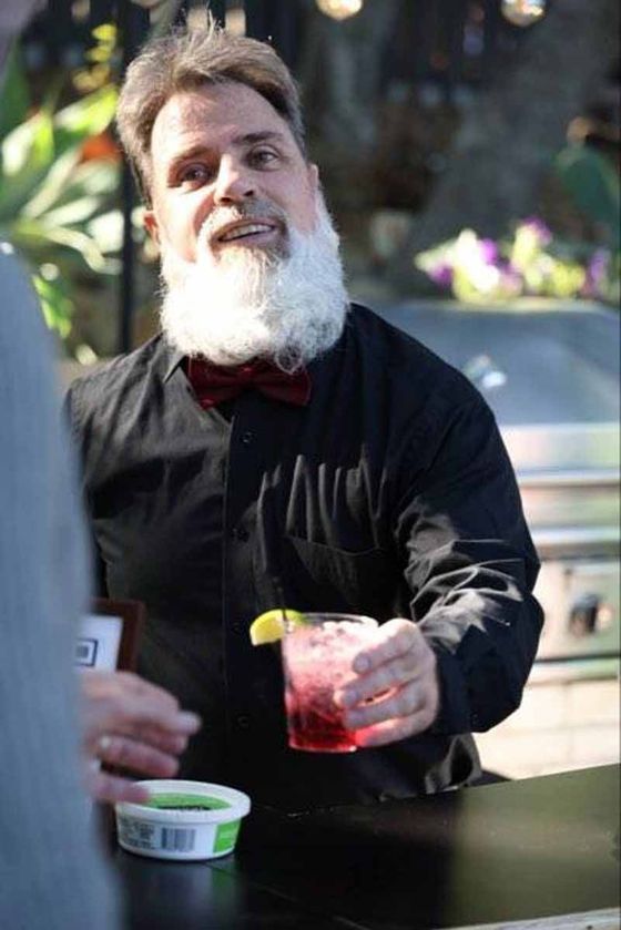 Man holding a drink beside a table outdoors, with a small tub in the foreground and blurred cars behind