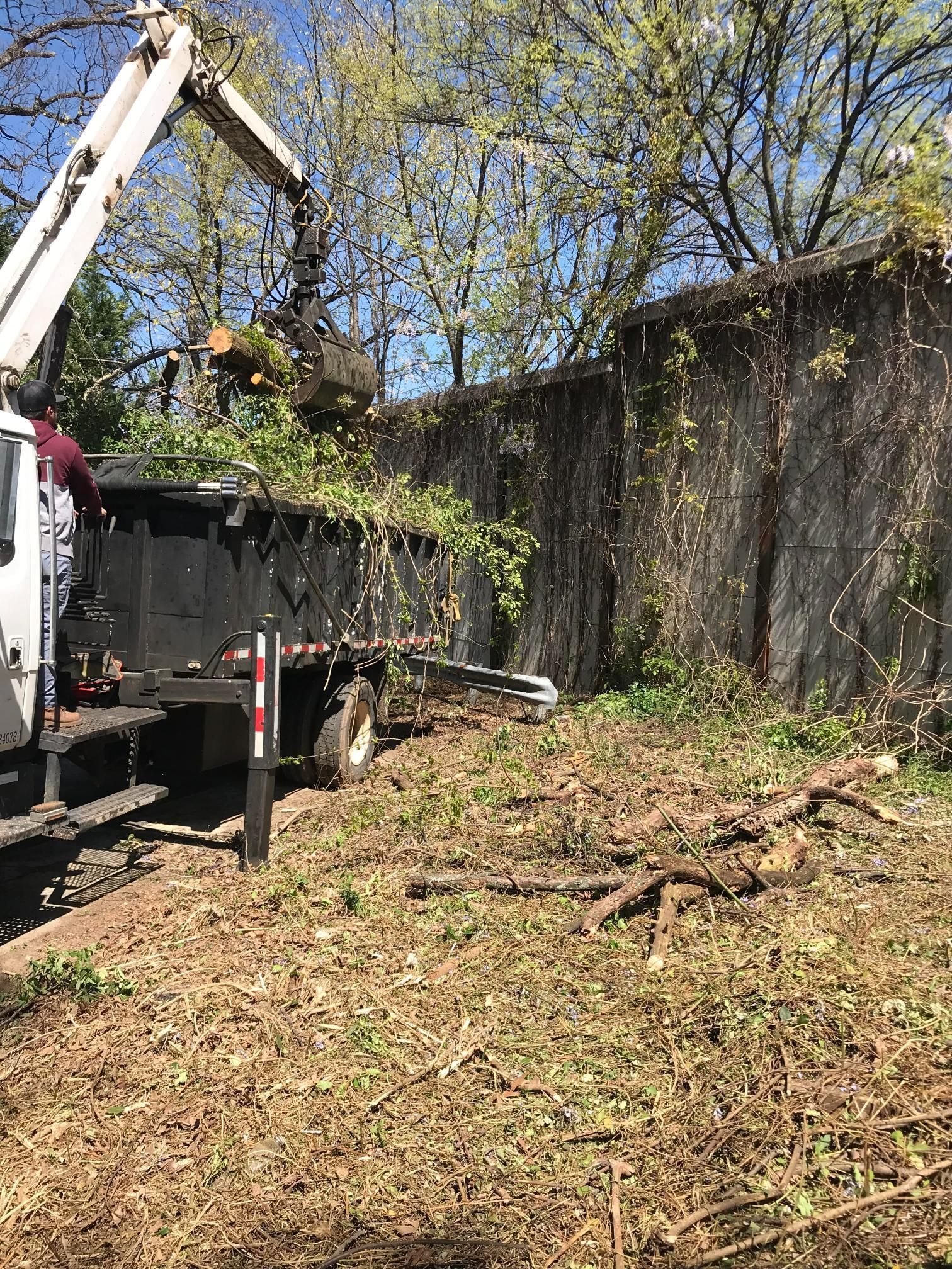 A truck with a hydraulic grapple crane lifting tree debris into its bed near a stone wall and wooded area.