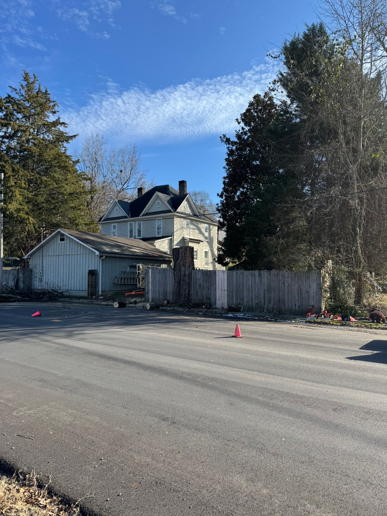A street view of a multi-story white house with a garage, adjacent trees, and a wooden fence under a clear blue sky.