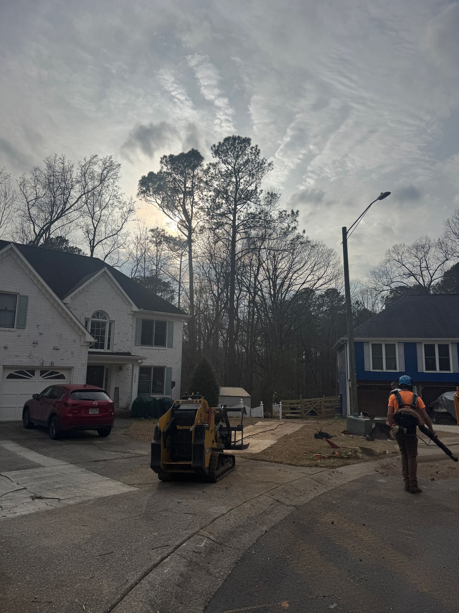 A person uses a leaf blower on a driveway near a yellow skid steer, construction debris, and two residential houses.