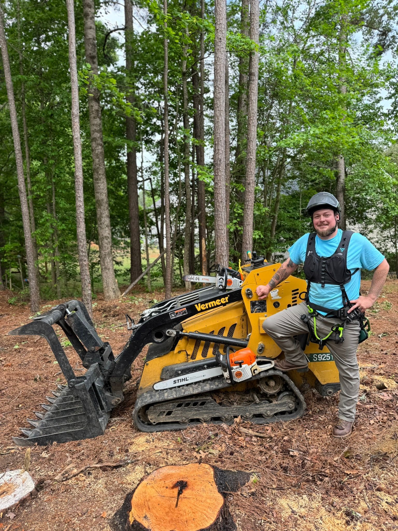 A smiling person in a blue shirt and safety gear stands next to a yellow tracked skid steer in a wooded area.