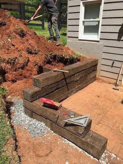 A person shovels a large dirt pile beside a house where a new wooden retaining wall is being constructed.