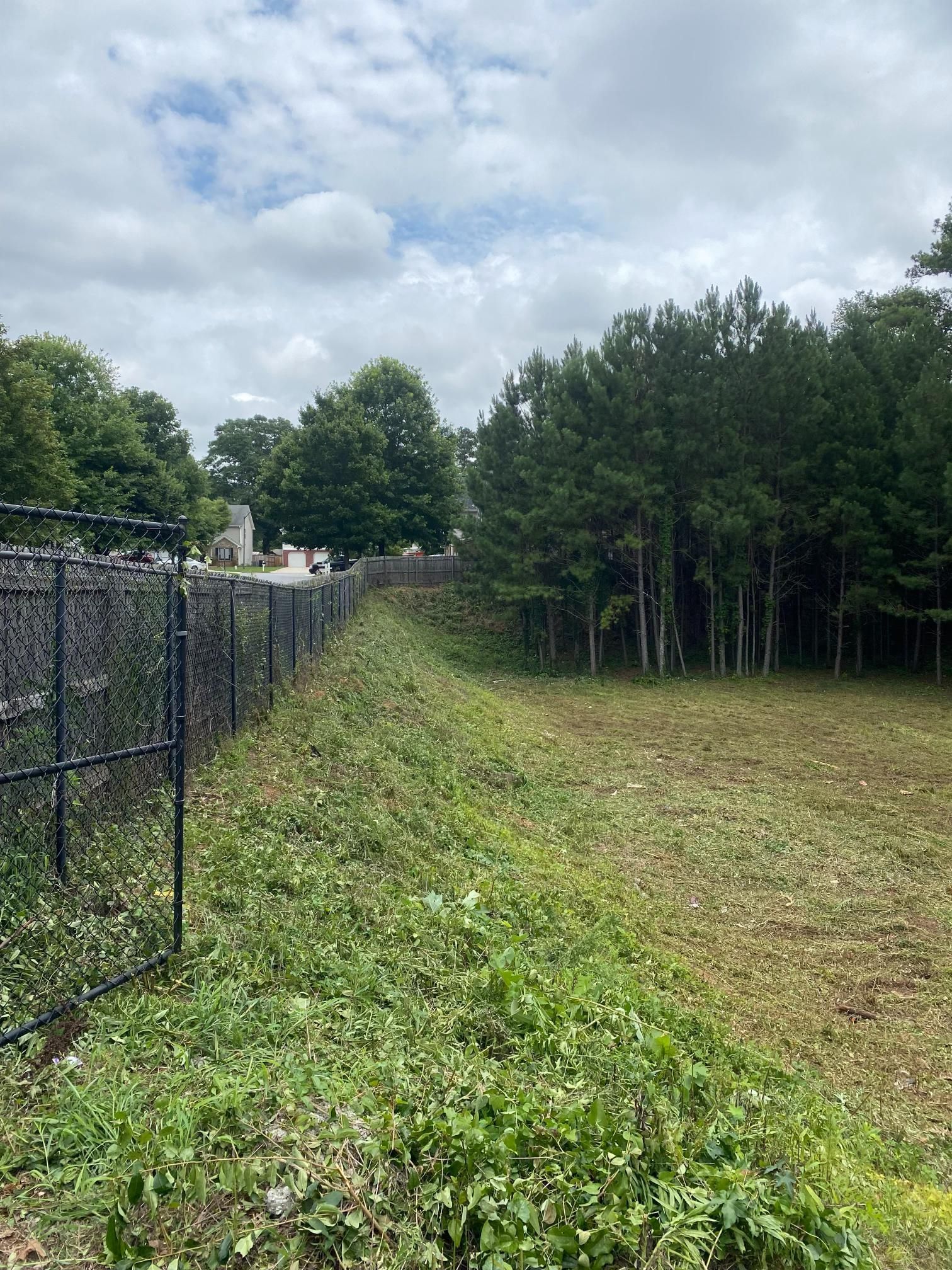 A chain-link fence runs along a grassy embankment next to a wooded area under a partly cloudy sky.