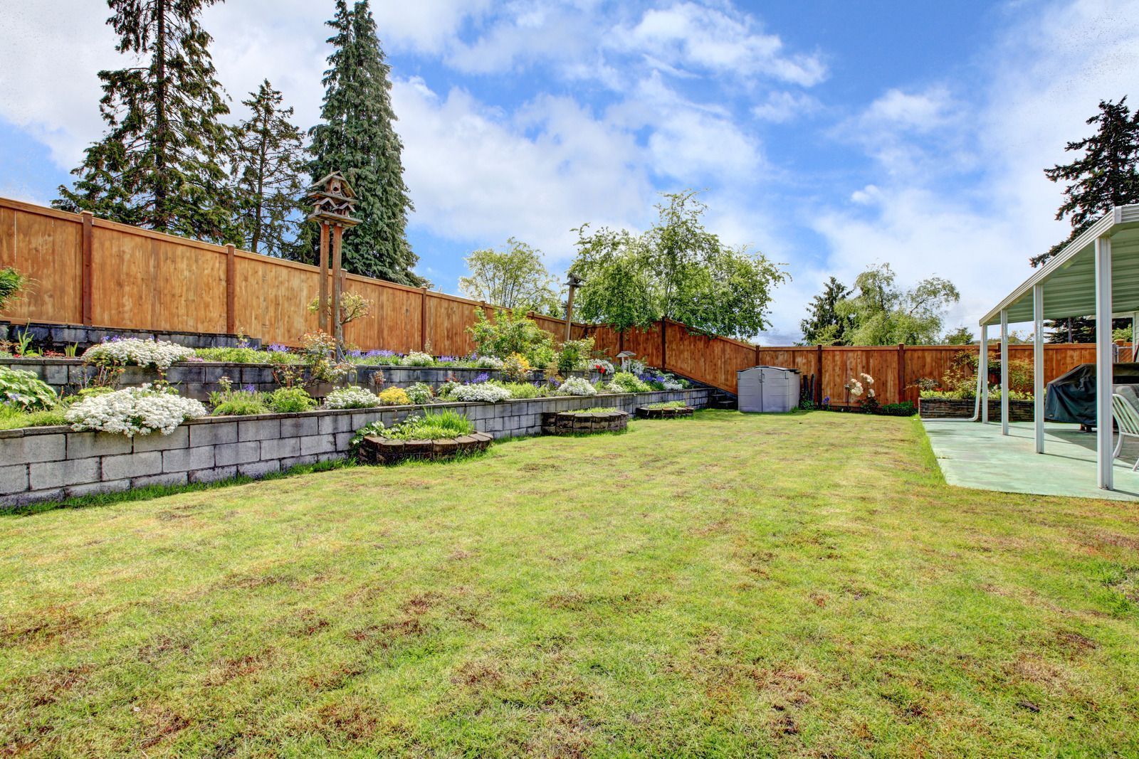 A green backyard with a tiered stone retaining wall garden, a wooden fence, and a patio cover under a blue cloudy sky.