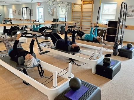 Three people practice Pilates on reformers in a light-filled studio with mirrors, wall bars, and equipment.