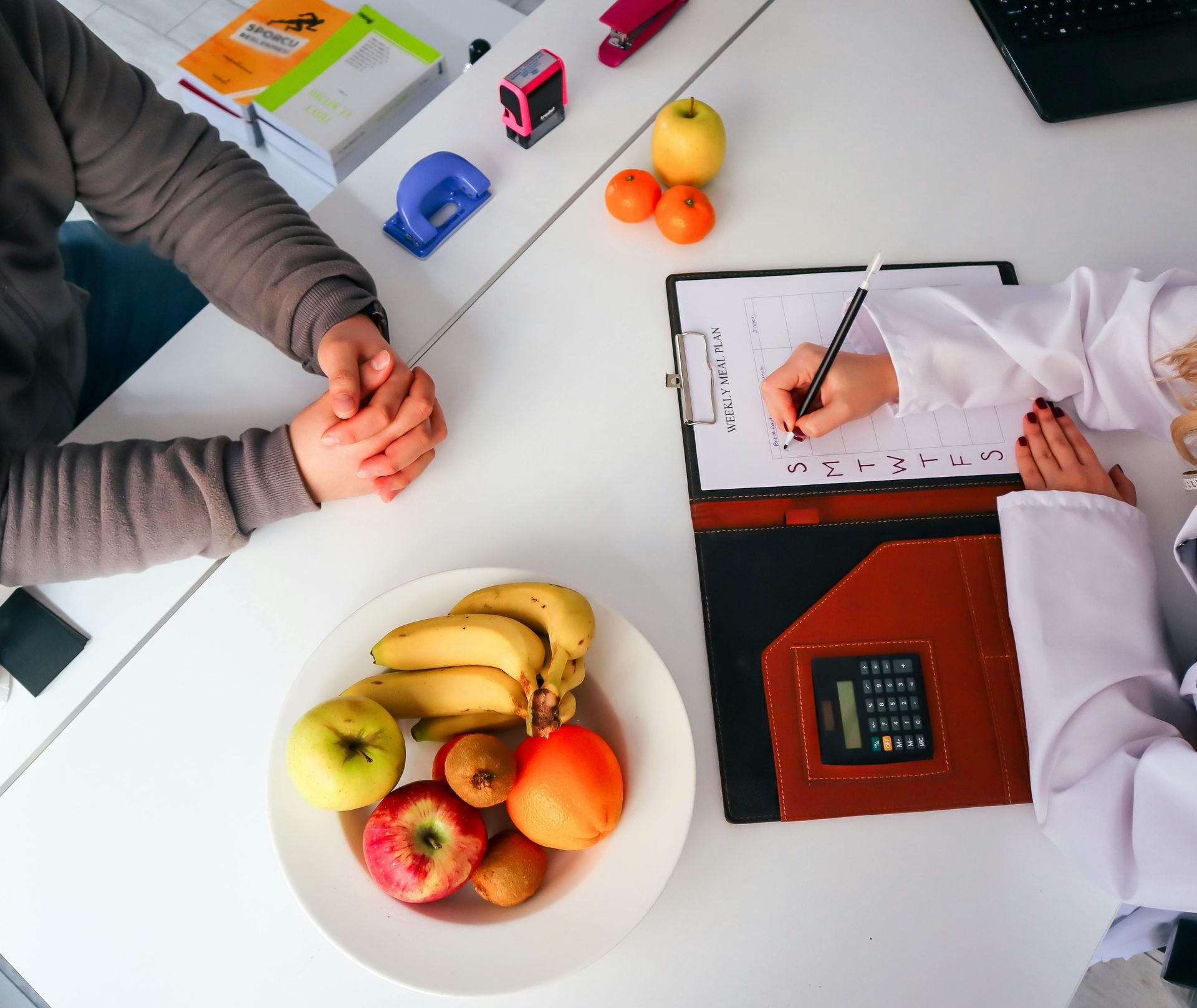 Two people discussing over a notebook at a desk with fruit, a calculator, and colorful toy cars.