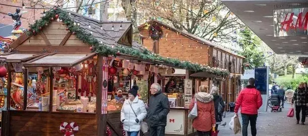 Christmas market with wooden stalls, people browsing, decorated with greenery and lights.