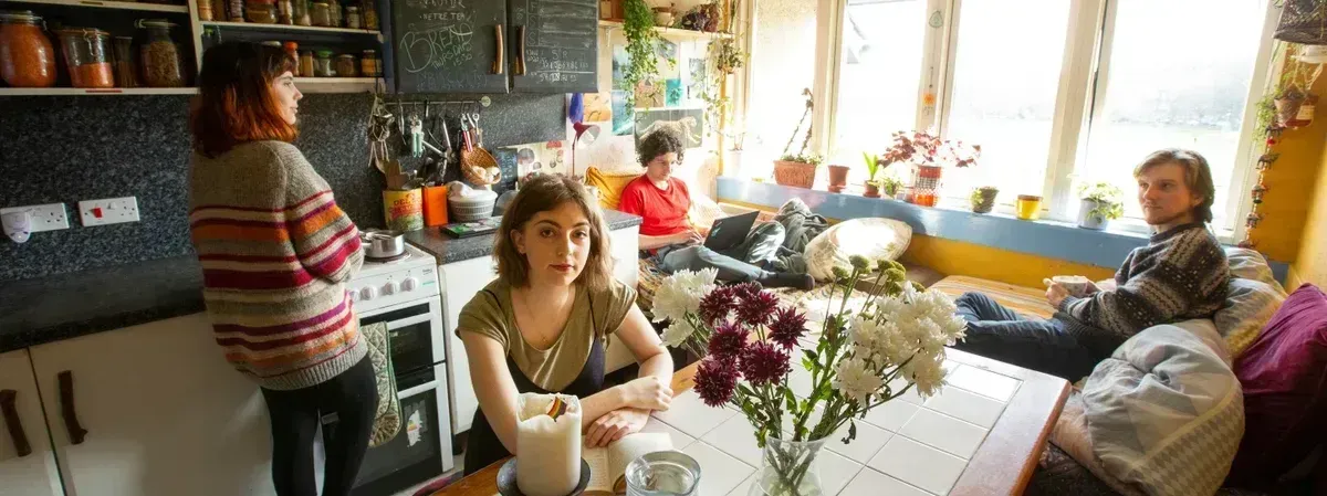 People gather in a kitchen with a bright window. A woman looks at the camera, flowers on the table.