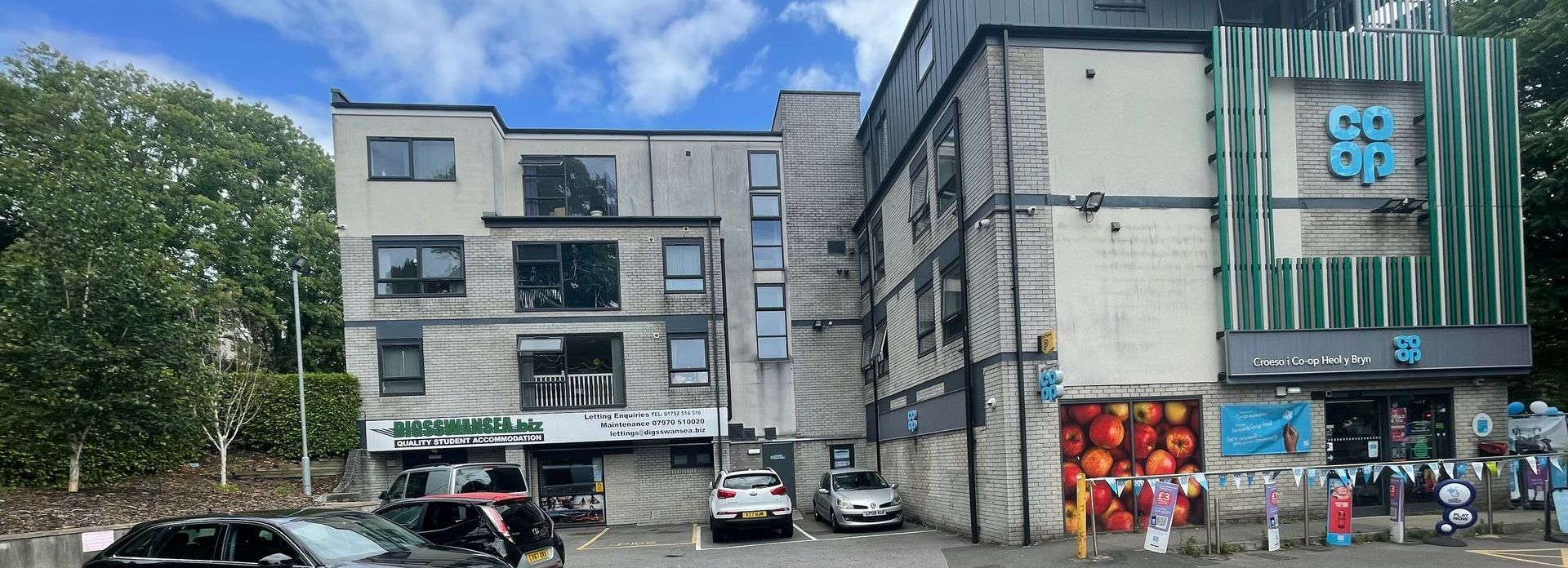 A multi-story building with a Co-op store, cars parked out front, and trees on the left.