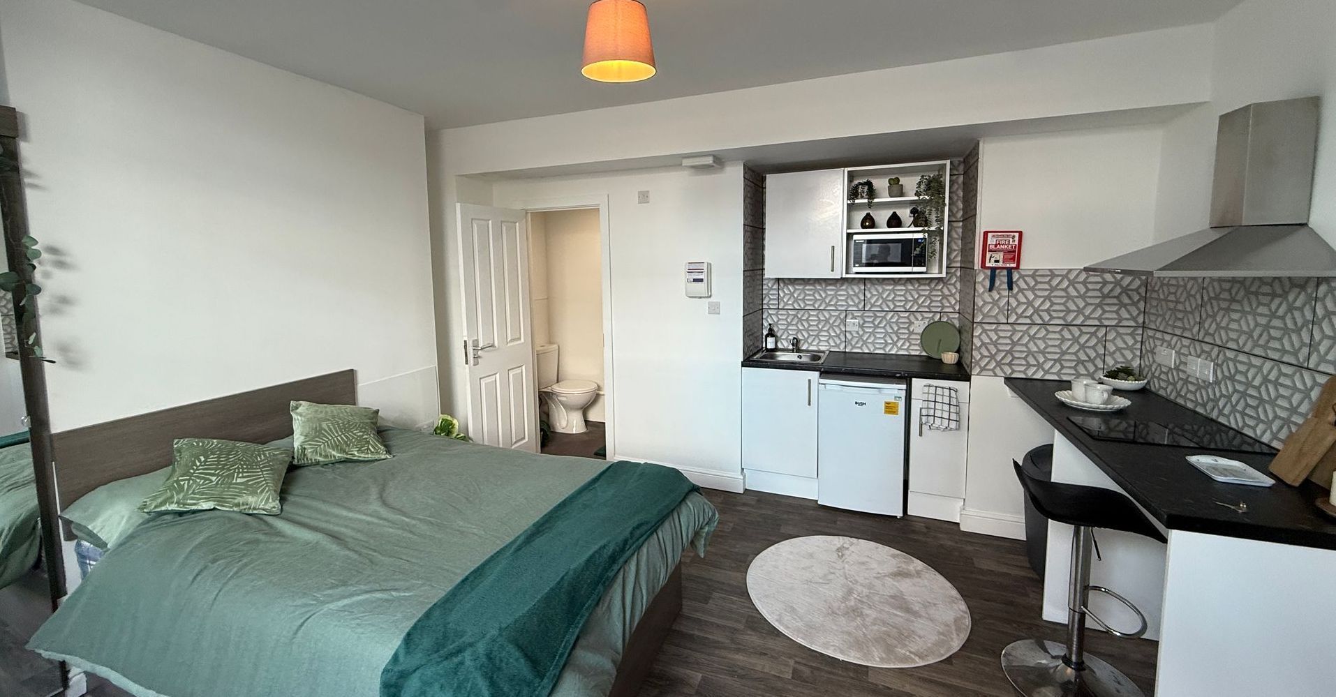 Studio apartment: bed, small kitchen with microwave, and bathroom visible. Green bedding, white walls.