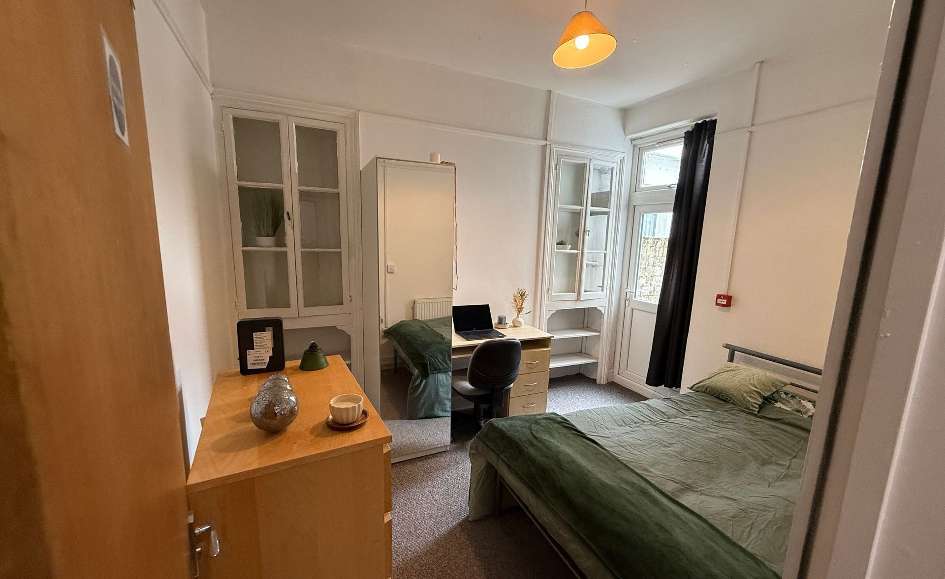 A small bedroom with bed, desk, wardrobe, and a dresser. White walls, beige furniture, and a window.