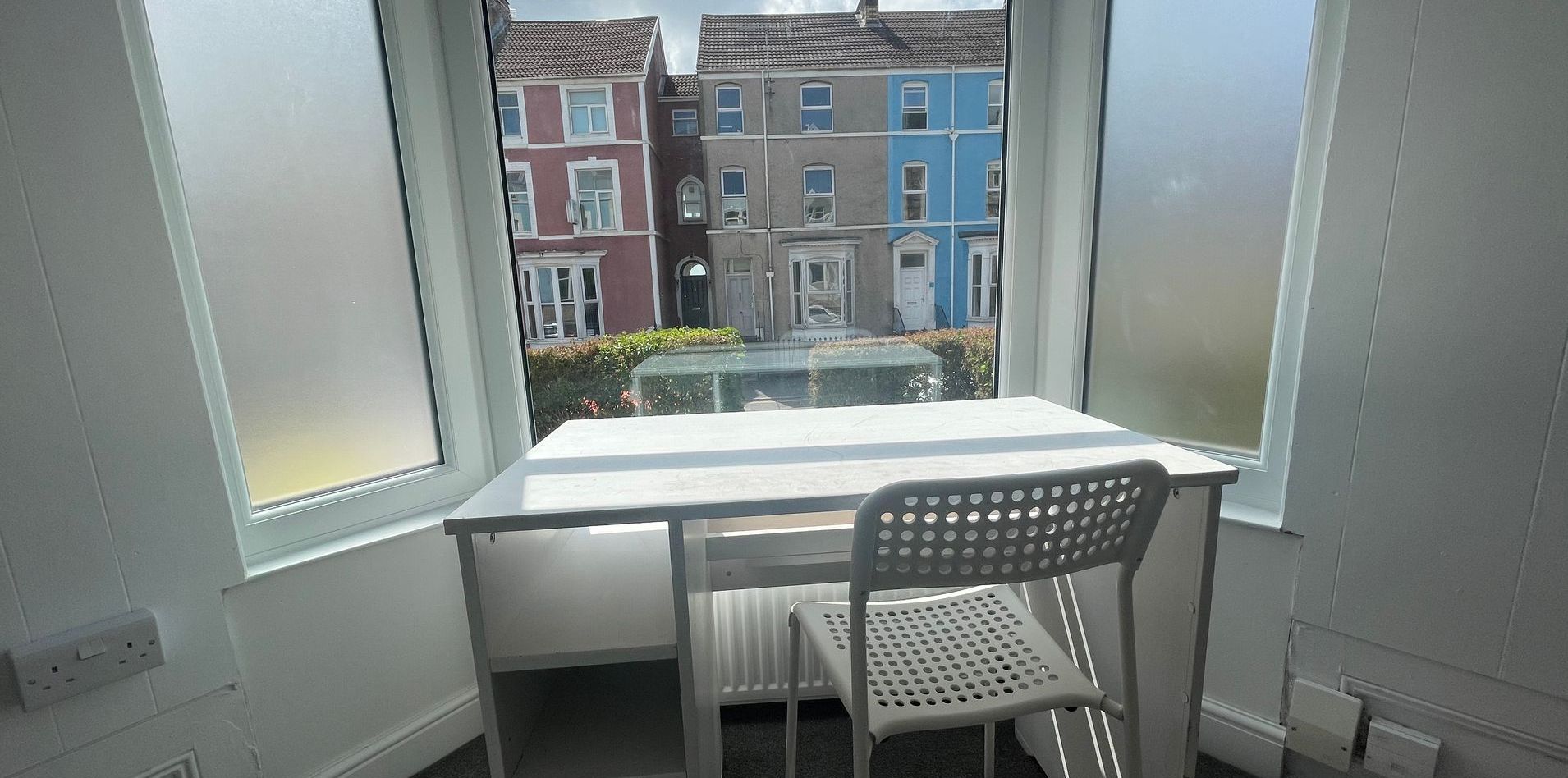 A small white desk and chair in front of a window overlooking colorful row houses.