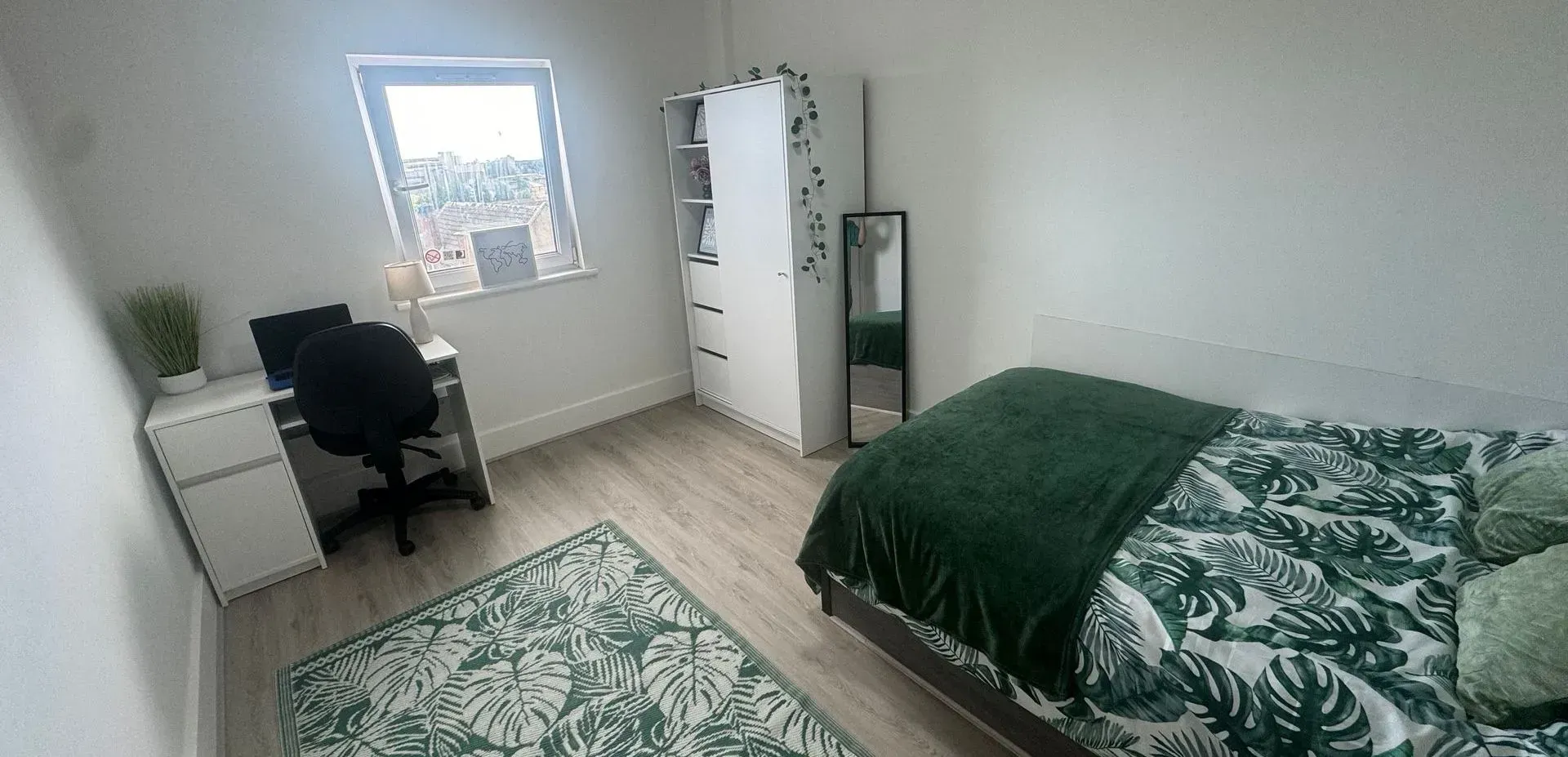 A bedroom with a bed, desk, rug, wardrobe, and mirror. The room has a green and white color scheme.