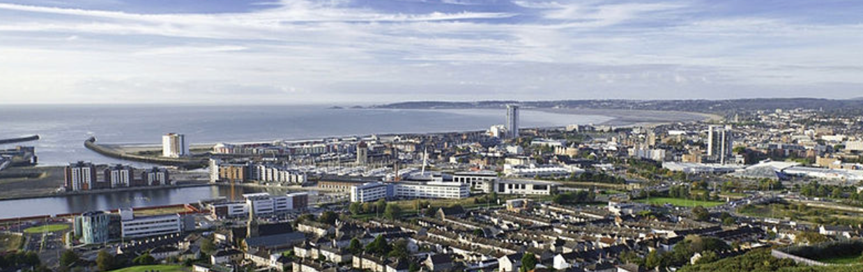 Aerial view of a city by the sea. Buildings and water are visible. The sky is blue.