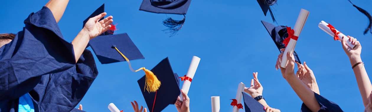 Graduates in blue gowns throwing caps and diplomas in the air against a clear blue sky.