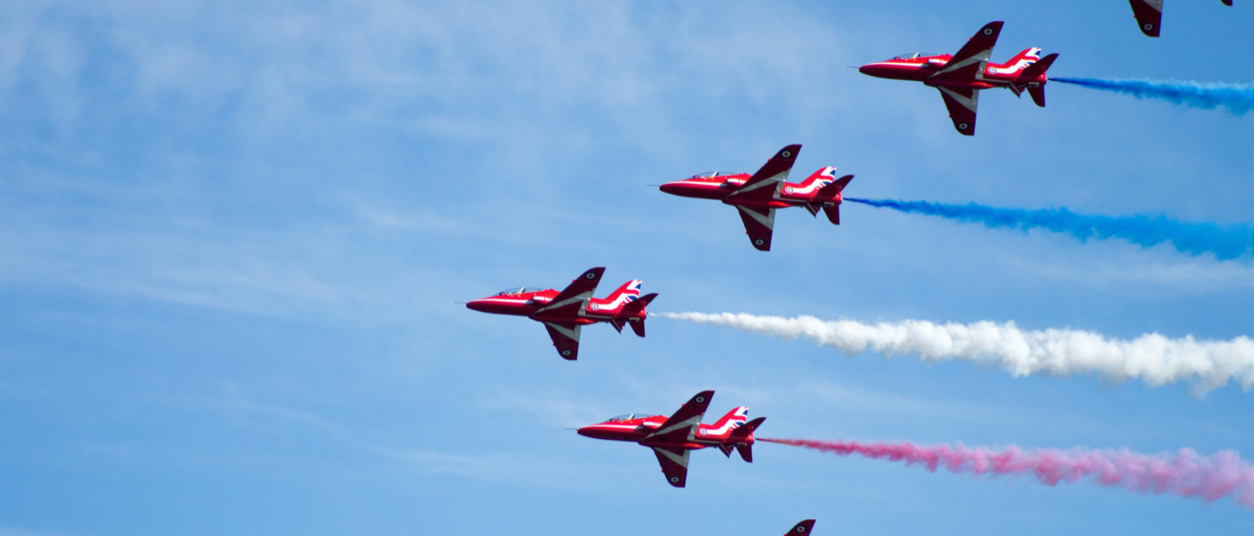 Red Arrows aerobatic team flying in formation against a blue sky, trailing red, white, and blue smoke.