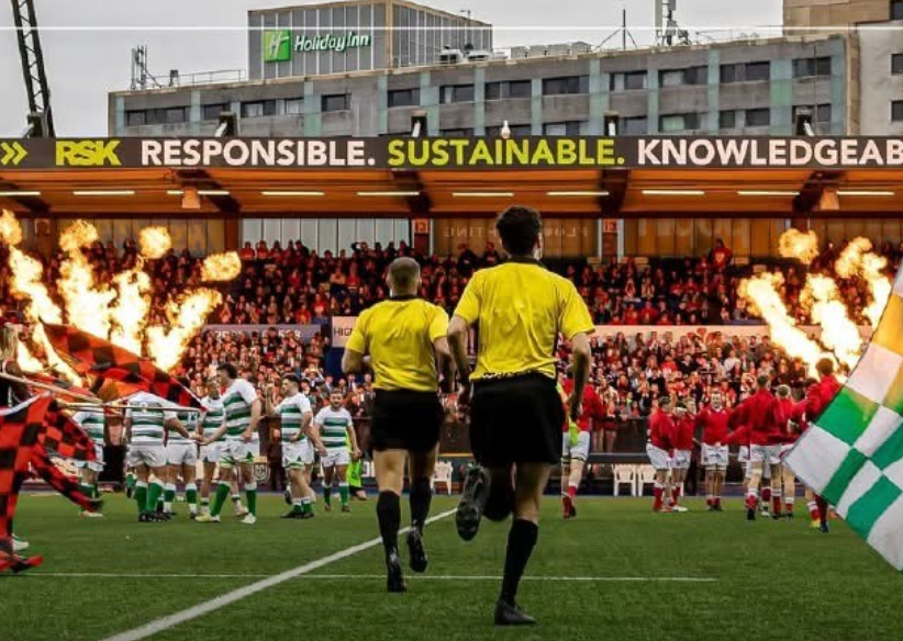 Two referees run onto a rugby field as a crowd watches with flames on either side.