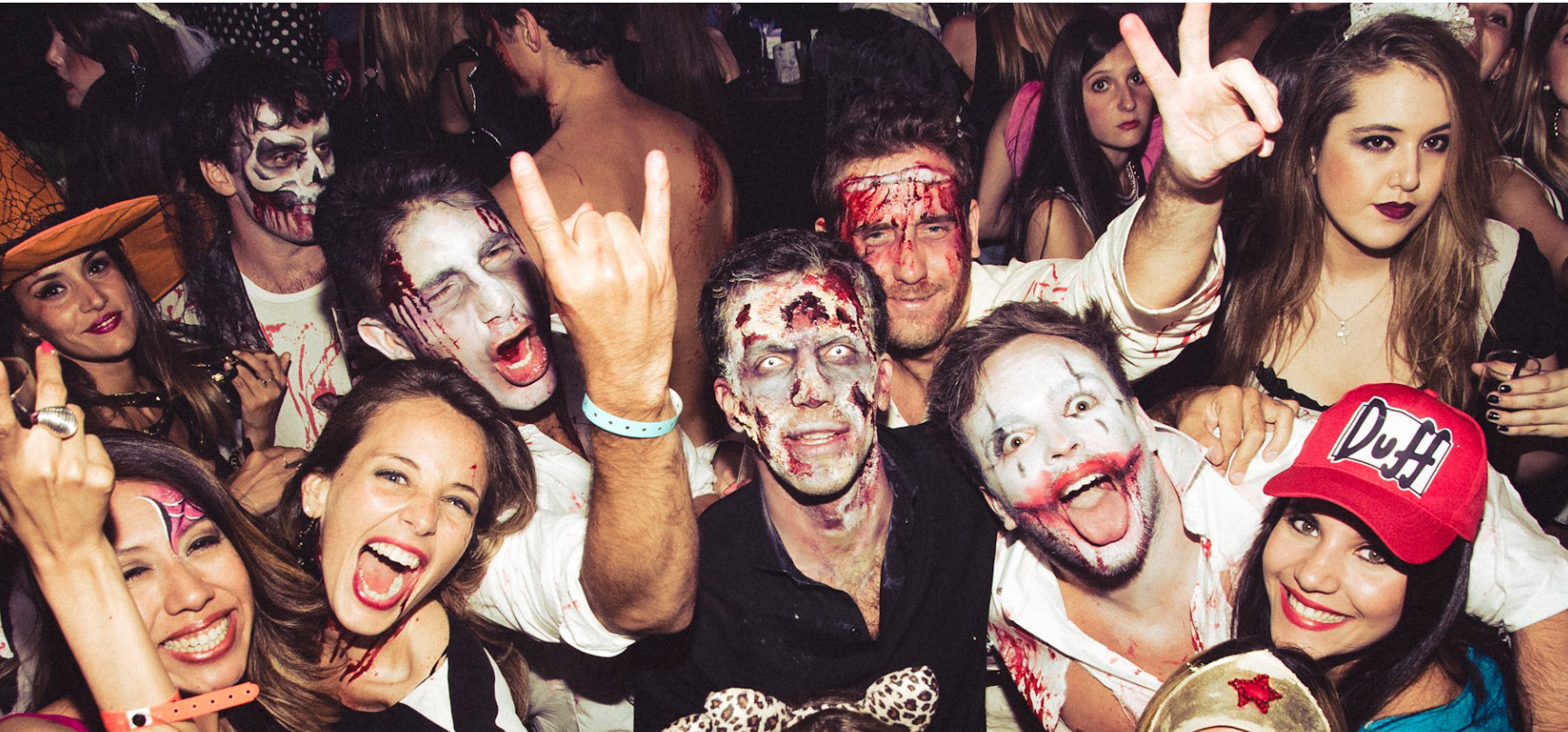 People in Halloween costumes at a party, some with zombie and clown makeup, raising hands, and smiling.