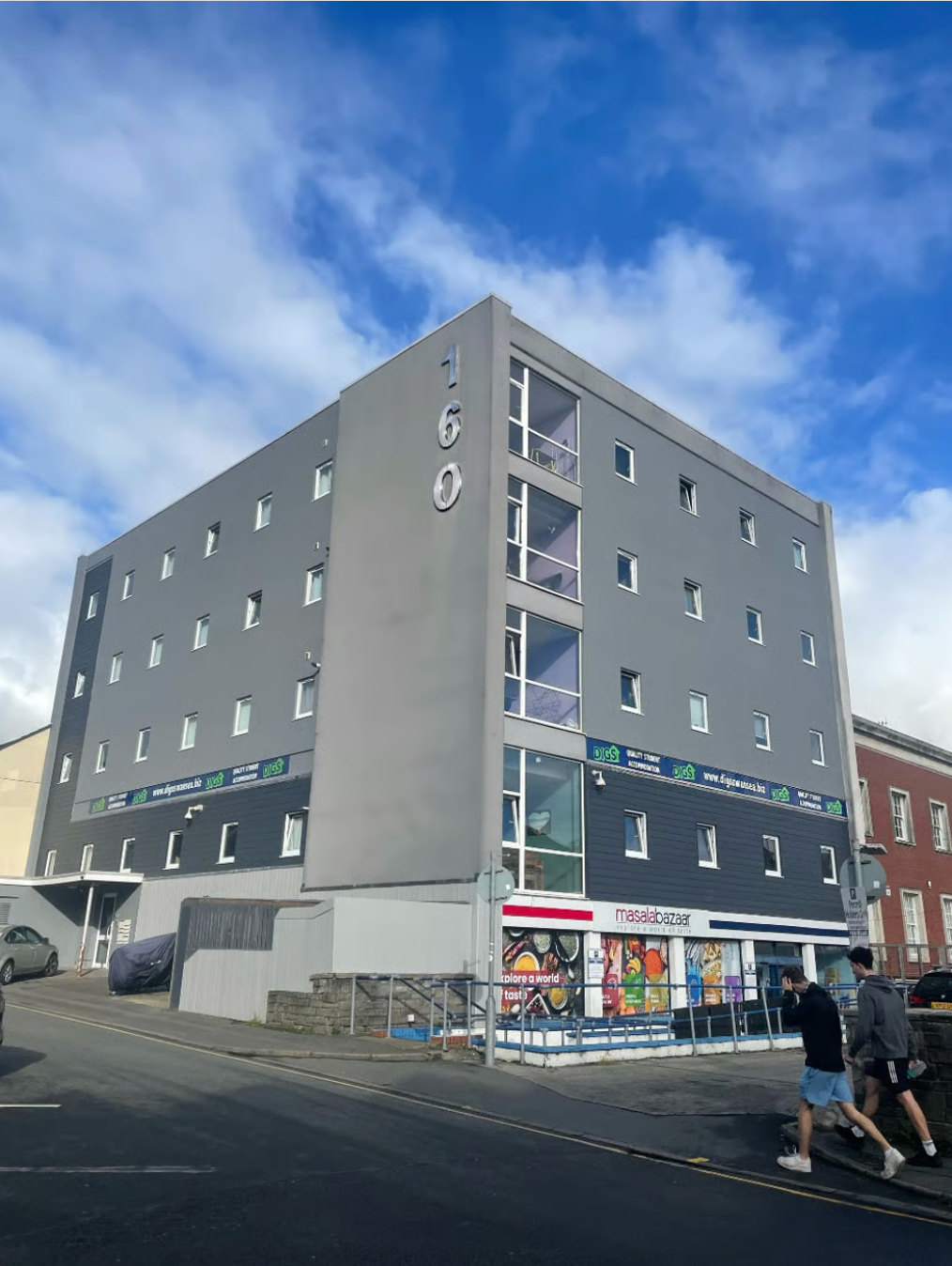 Gray multi-story building with small windows. A shop is on the ground floor, people walking nearby. Blue sky.