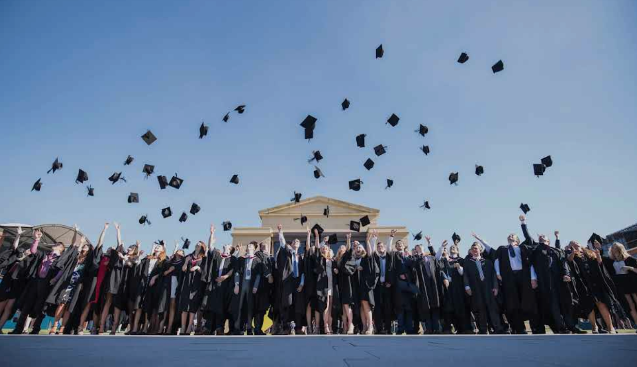 Graduates in black gowns toss mortarboards into the air in front of a building with a blue sky background.