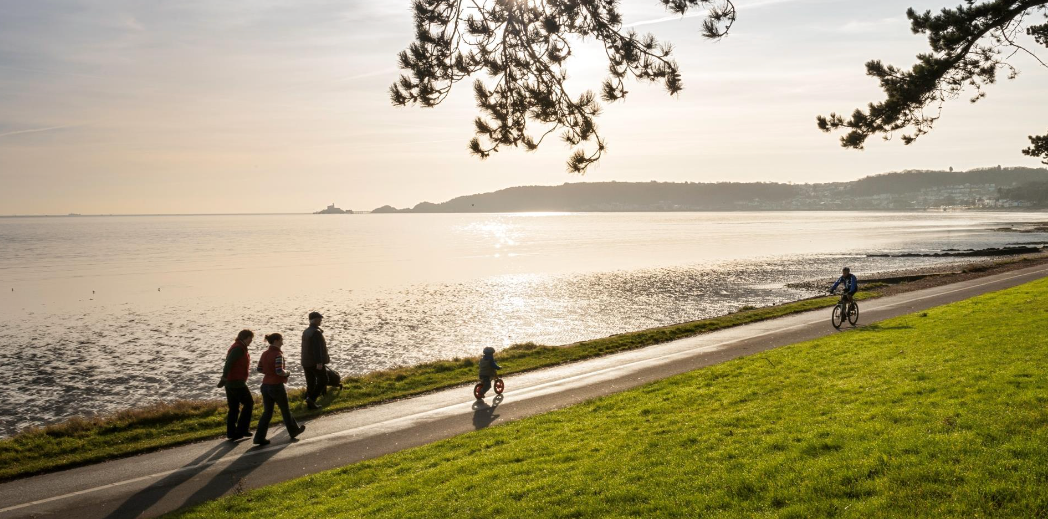 People walk and bike along a seaside path at sunrise. The water reflects the golden light.
